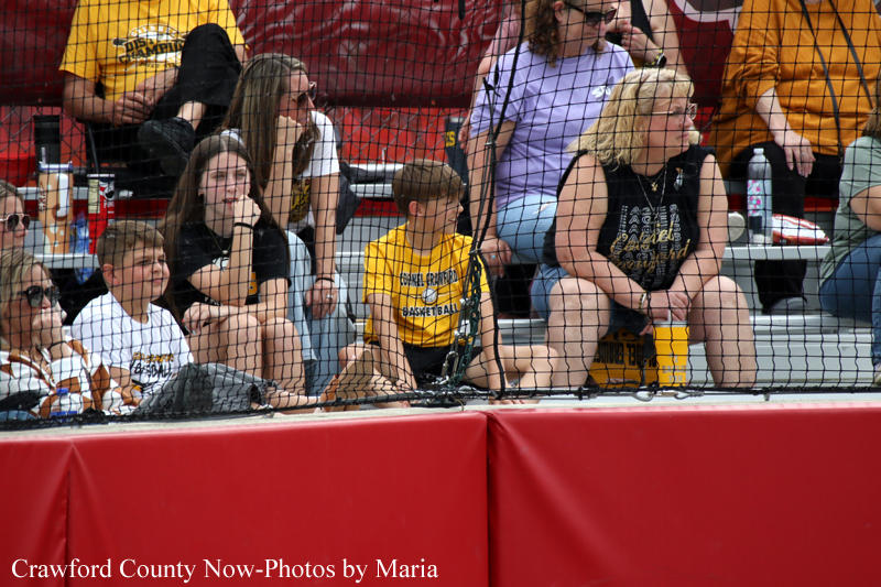Group of spectators behind a protective net watching a basketball game, with several kids in yellow shirts seated in the front row.