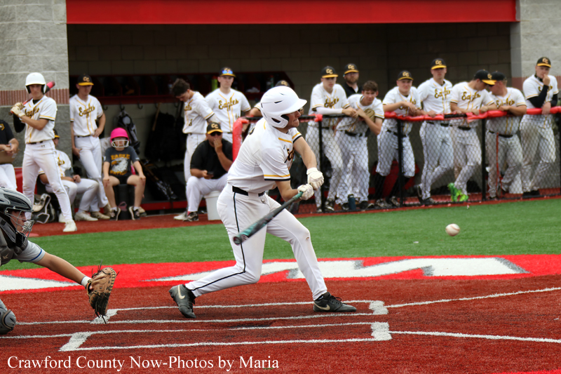 Baseball batter in white uniform swings at a pitch near home plate as teammates watch from the dugout behind him.