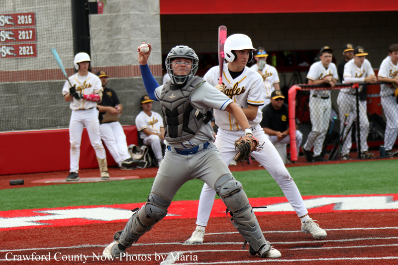 Baseball catcher in full protective gear at home plate, preparing to throw, with a batter in white uniform behind him and teammates in the dugout in the background.