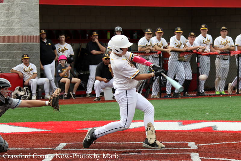 Baseball batter in a white uniform swings at a pitch at home plate with teammates watching from the dugout in the background.