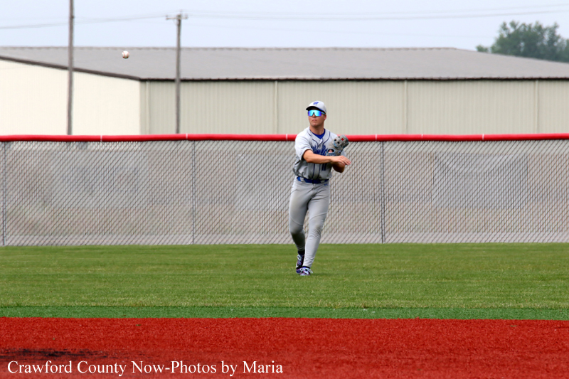 Baseball fielder in gray uniform stands on the outfield grass, glove ready as a ball flies overhead near a chain-link fence and red warning track.