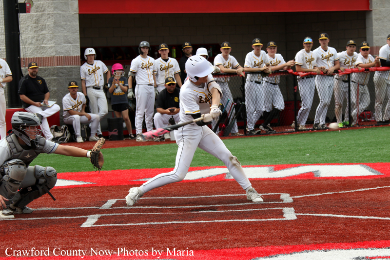 Baseball batter in white swings at a pitch, with catcher crouched behind home plate and teammates watching from the dugout.