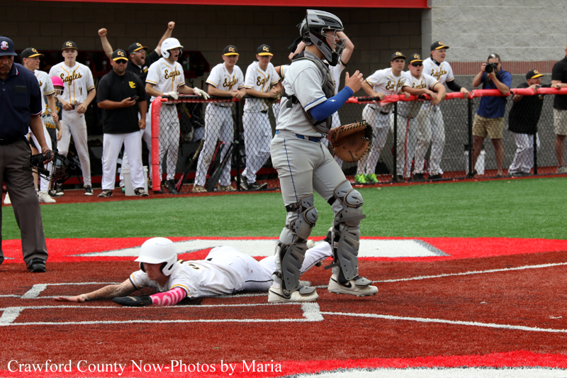Baseball at home plate: a catcher in gray protective gear stands as a sliding runner in white lunges toward the plate, with the dugout and crowd in the background.