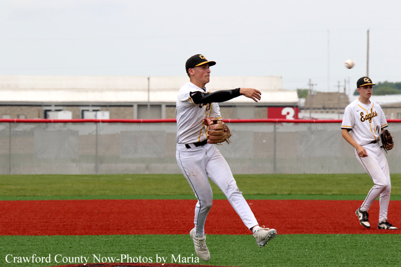 Baseball pitcher in white uniform mid-windup throwing a pitch on a red-dirt infield, a teammate watches in the background.