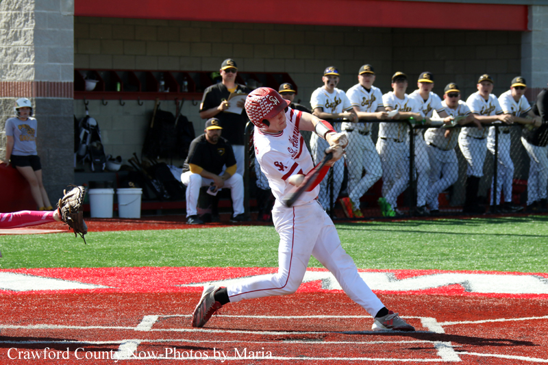 Baseball batter swinging at a pitch in a red turf-surface batter's box with teammates watching from the dugout behind him.