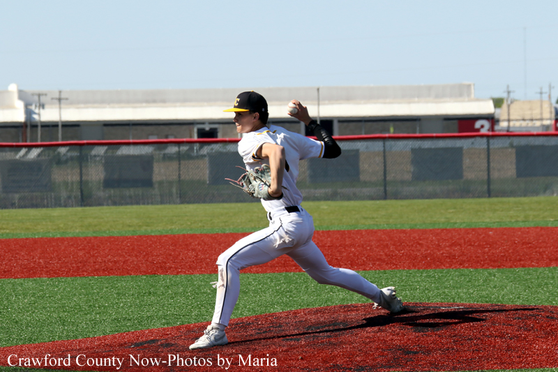 Baseball pitcher in white uniform throws from the mound on a red-dirt infield with green outfield, fence and stands in the background.