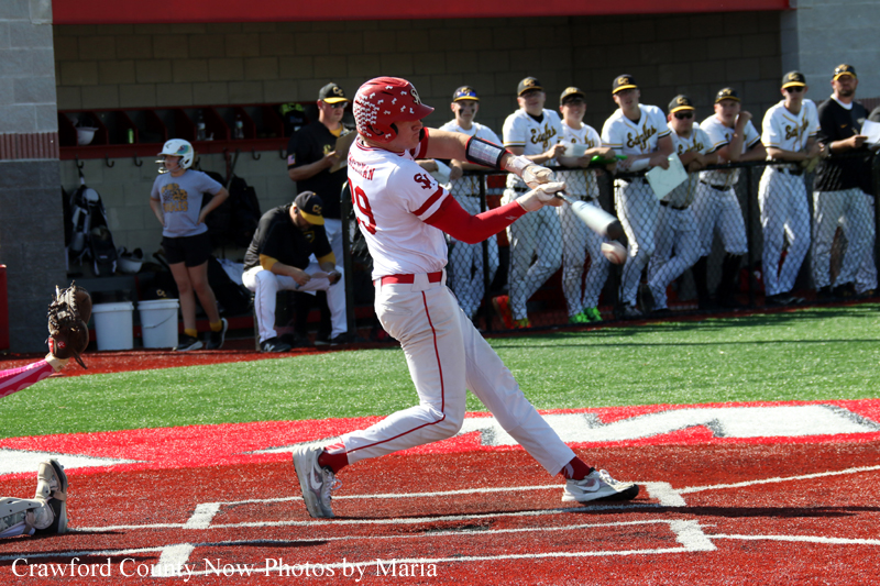 Baseball batter in white and red swings at a pitch as teammates watch from the dugout in the background.