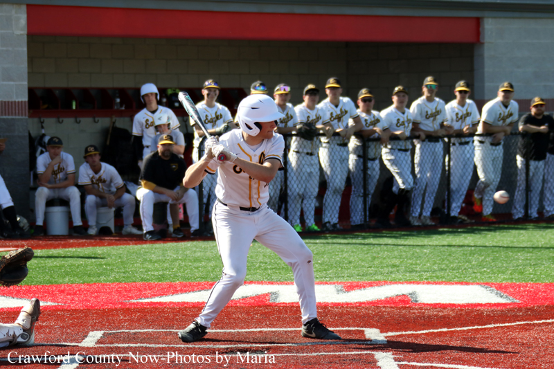 Baseball batter in a white uniform at home plate, poised to swing as teammates watch from the dugout behind him.