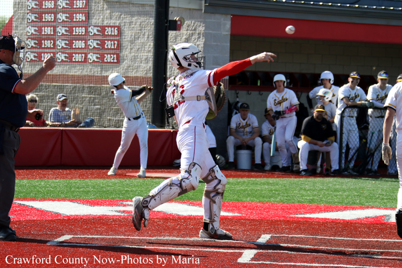 Baseball catcher in white gear steps toward home plate after a play, with teammates watching from the dugout.