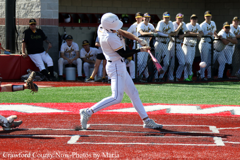 Baseball batter in white uniform swings at a pitch at home plate while teammates watch from the dugout in the background.