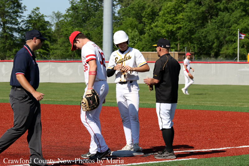 Baseball players gather at first base on a sunny field; one Eagles player adjusts gear while an umpire and a coach stand nearby.