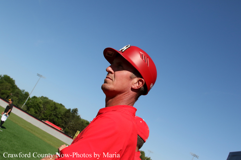 Close-up of a man wearing a red helmet and red jersey, looking up against a clear blue sky at a baseball field.