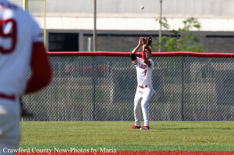 Baseball outfielder in white uniform catching a high pop foul near the fence, glove raised above his head as the ball drops in. A teammate is blurred in the foreground on the left.