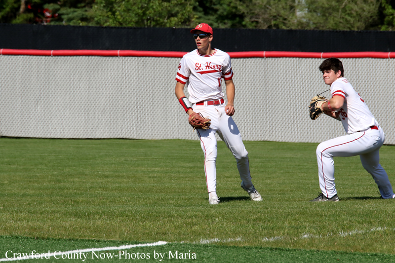 Two baseball players in white and red uniforms on a grass field; one stands with a glove ready while the other crouches in a fielding stance near a chain-link fence in the background.