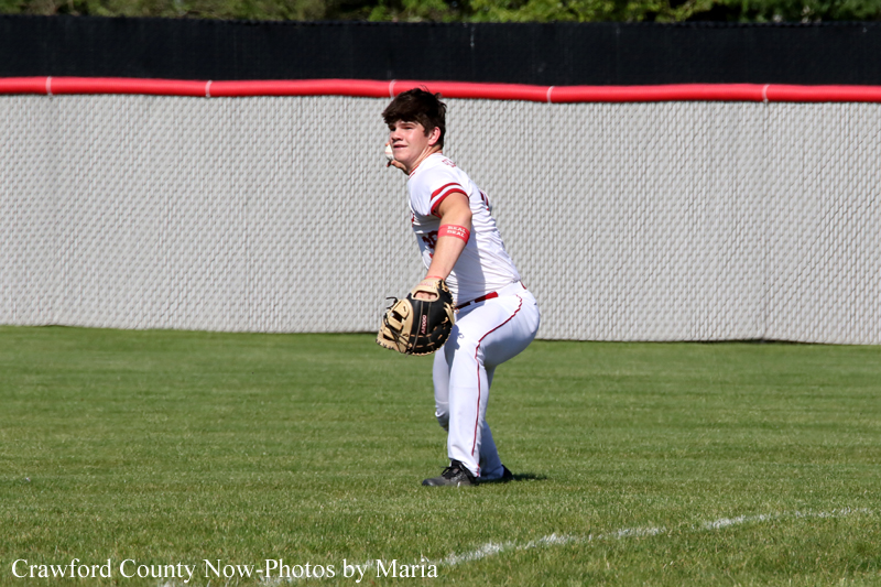 Baseball pitcher in a white uniform with red piping, glove ready as he prepares to throw on a grassy field with a chain-link fence behind him