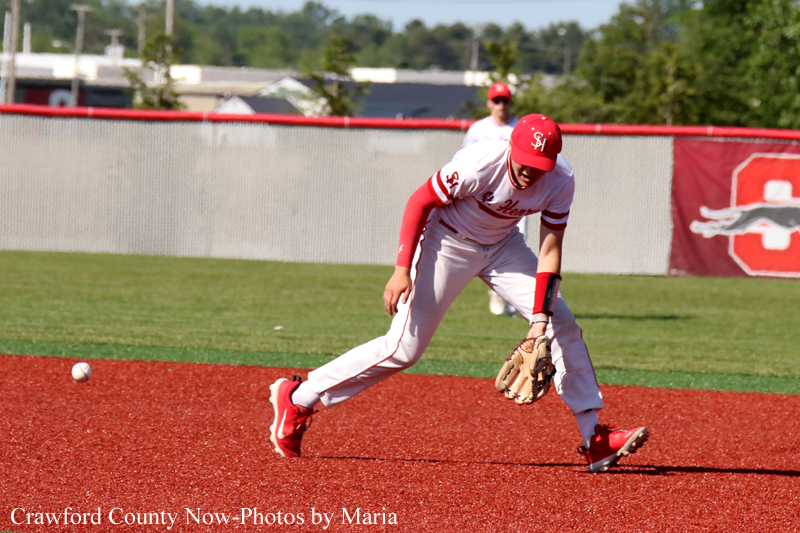 Baseball infielder in red and white uniform stretches to field a ground ball on a red dirt infield.