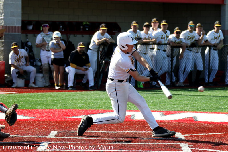 Baseball batter in white uniform swings at a pitch on a red infield, with teammates watching from the dugout in the background.