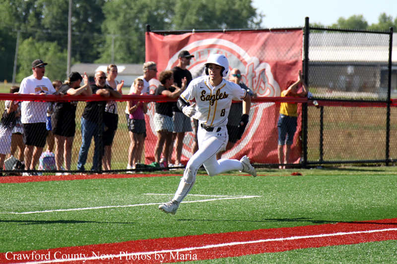 Female softball player in a white Eagles uniform sprints along the baselines while crowd watches from behind a red fence and banner in a stadium.