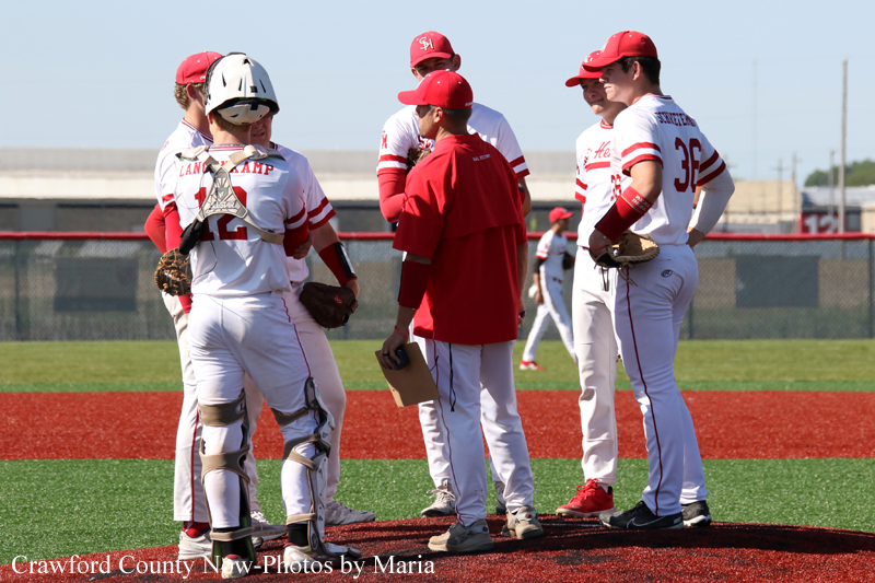 Group of baseball players in white and red uniforms listening to a coach on a baseball diamond.