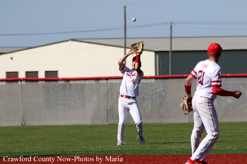 Baseball players in white and red uniforms on a grassy field; a fielder reaches up with his glove to catch a high ball, another player stands nearby near the infield fence.