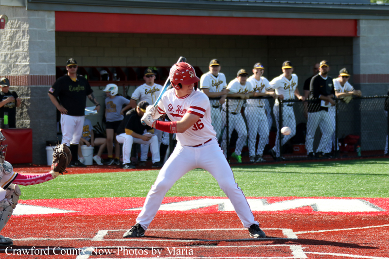 Baseball batter in white and red swings at a pitch on a red-dirt field, with teammates watching from the dugout behind him.