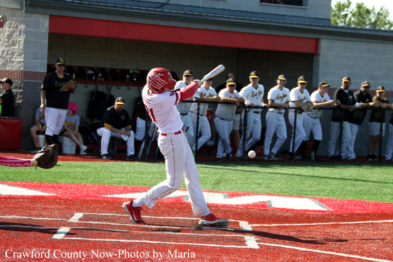 Baseball batter in white uniform swings at a pitch near home plate with a red helmet and bat, while teammates watch from the dugout in the background.