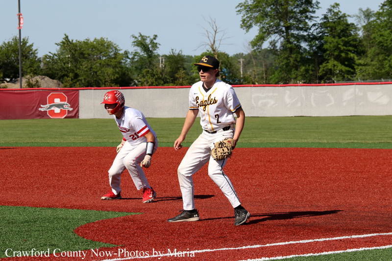 Two youth baseball players on a red infield, one in a white 'Eagles' uniform with glove ready at first, the other in red helmet nearby as a baserunner approaches behind him.