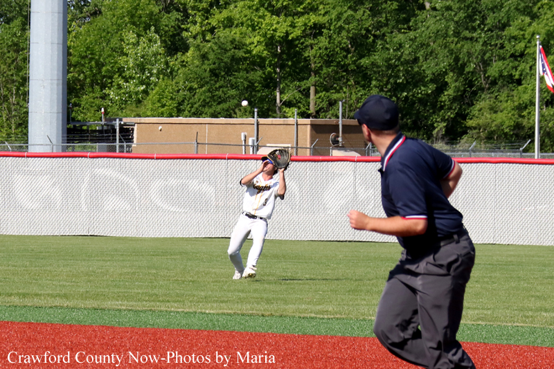 Young baseball player in white uniform catches a fly ball near the outfield fence with an umpire running in foreground at a sunny field.