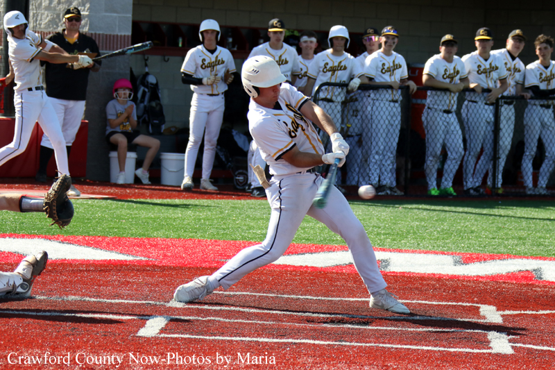 Baseball batter in white uniform swings at a pitch at home plate, with teammates watching from the dugout behind the fence.