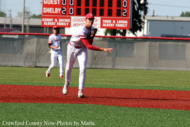 Baseball pitcher in white and red winds up on the mound as a teammate watches, with a scoreboard visible in the background.