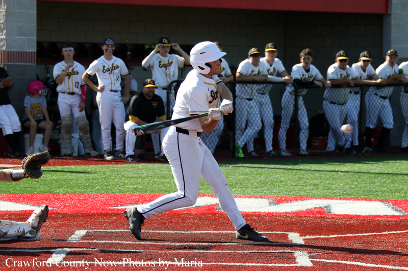 Baseball batter in a white uniform swings at a pitch on a red infield as teammates watch from the dugout behind a chain-link fence.