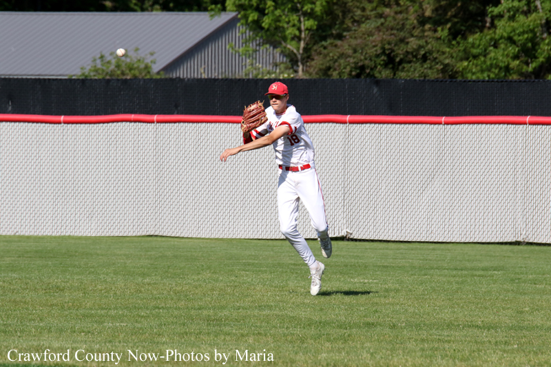 Baseball pitcher in white uniform with red cap throws a ball on a green outfield.