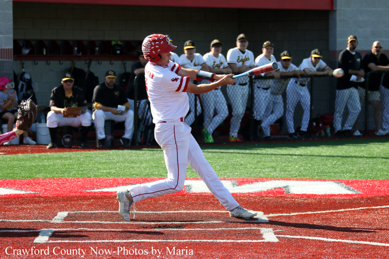 Baseball batter in white and red swings at a pitch during a game, with teammates in the dugout watching in the background.