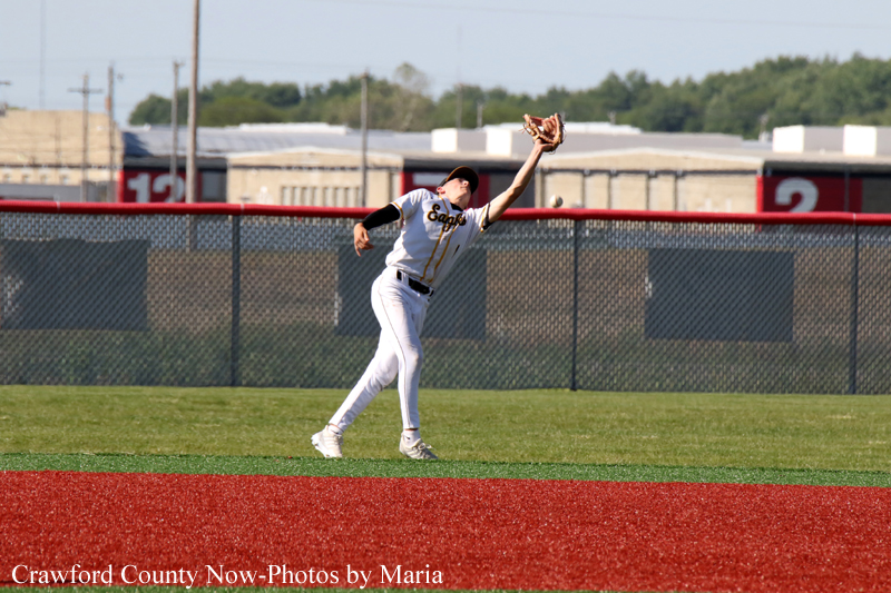 Baseball player in a white uniform mid-throw on a green field, glove raised, with a chain-link fence and red dirt infield in the background.