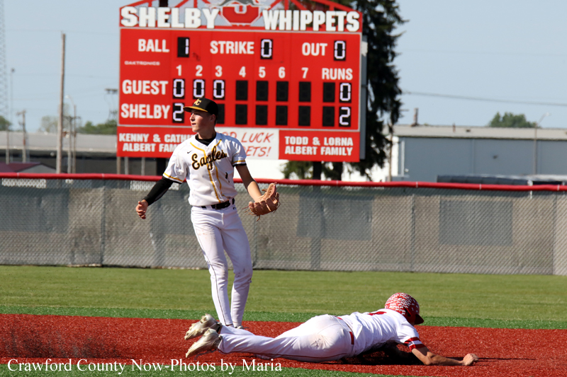 Baseball field moment: a fielder in white and gold stands at a base while a runner slides headfirst on the red dirt infield, with a large scoreboard in the background.