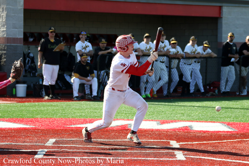 Baseball player in white and red uniform swings at a pitch at home plate, with teammates and spectators in the dugout area behind him.