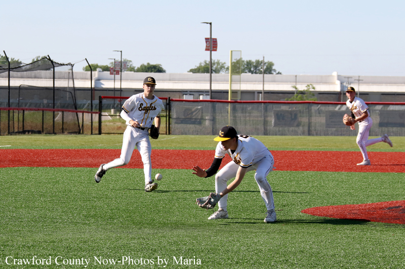 Baseball game on artificial turf as a fielder lunges for a ground ball while teammates watch nearby on the red infield.