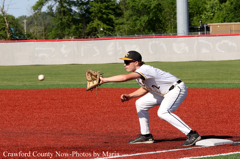 Baseball infielder in white uniform catches a fast ground ball near first base on a red dirt infield, glove stretched forward.