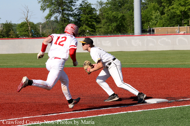 Baseball runner in white with red number 12 sprints toward a bag as the fielder in white crouches with a glove at the base to tag him out.