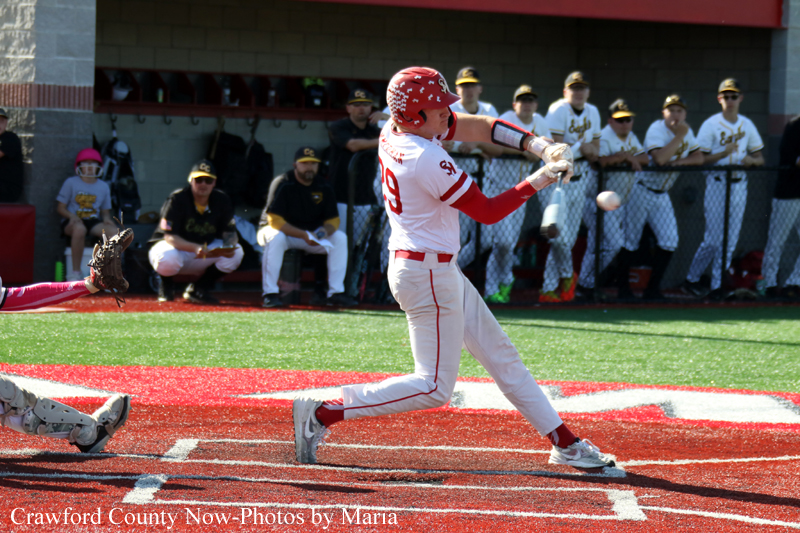 Baseball batter in a white and red uniform swings at a pitch on a red infield while teammates watch from the dugout in the background.