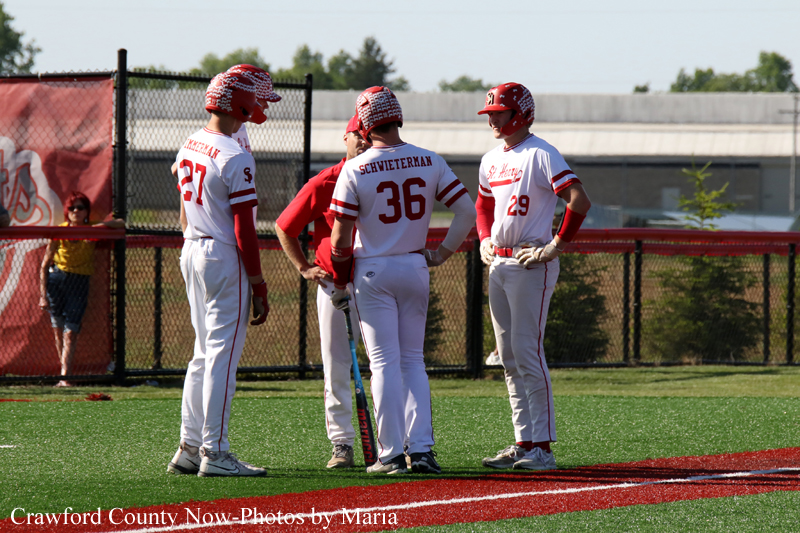 Three baseball players in white and red uniforms stand in a huddle on the field, chatting near the on-deck area.
