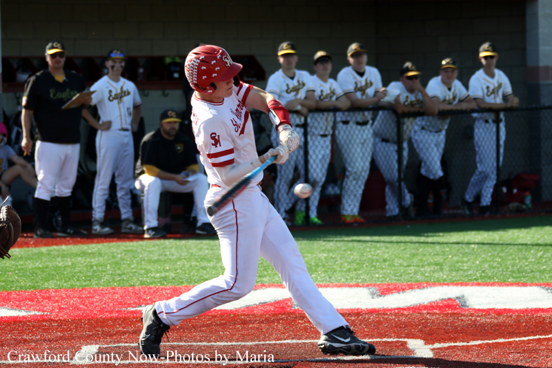 Right-handed batter in a white uniform and red helmet swings at a pitch at home plate with teammates watching from the dugout behind a chain-link fence.