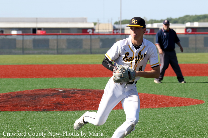 Baseball pitcher in white Eagle uniform on the mound preparing to throw, with an umpire in the background on a sunny field.