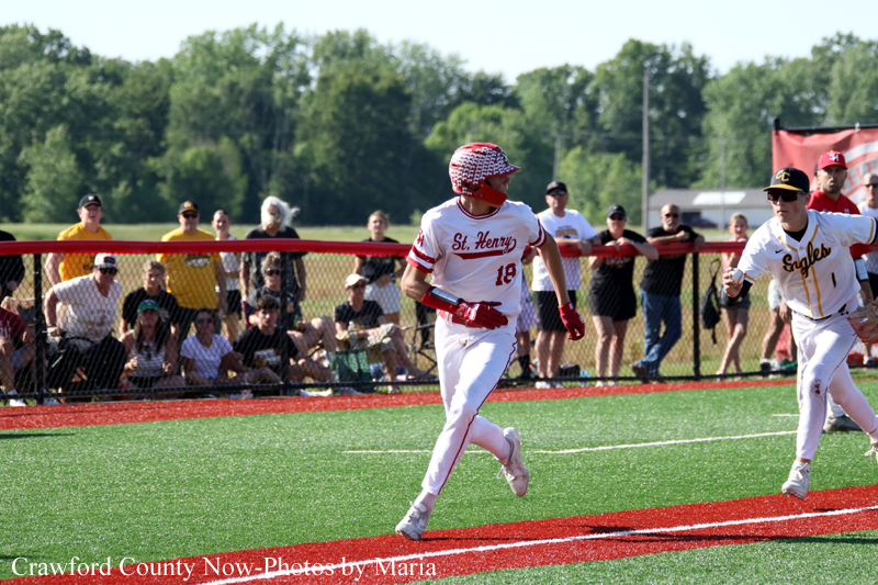 Baseball player in a St. Henry uniform runs on a red turf field as spectators watch from behind a chain-link fence.