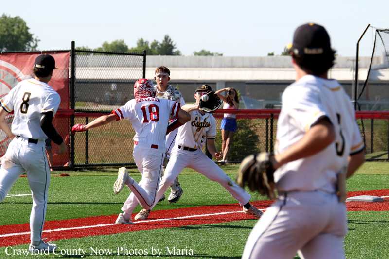 Baseball game action: a runner slides into home as a fielder reaches to tag him, with other players and a fence in the background.