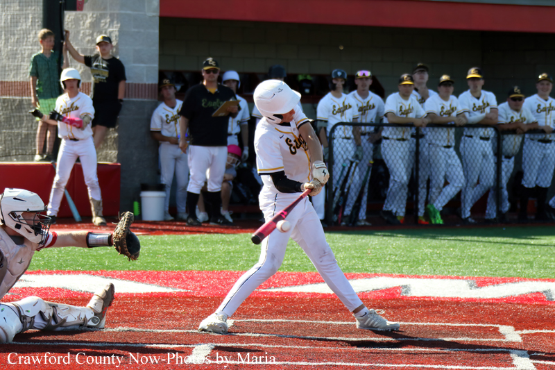 Baseball batter in white uniform swings at a pitch as the catcher dives to stop the ball, with teammates watching from the dugout in the background.