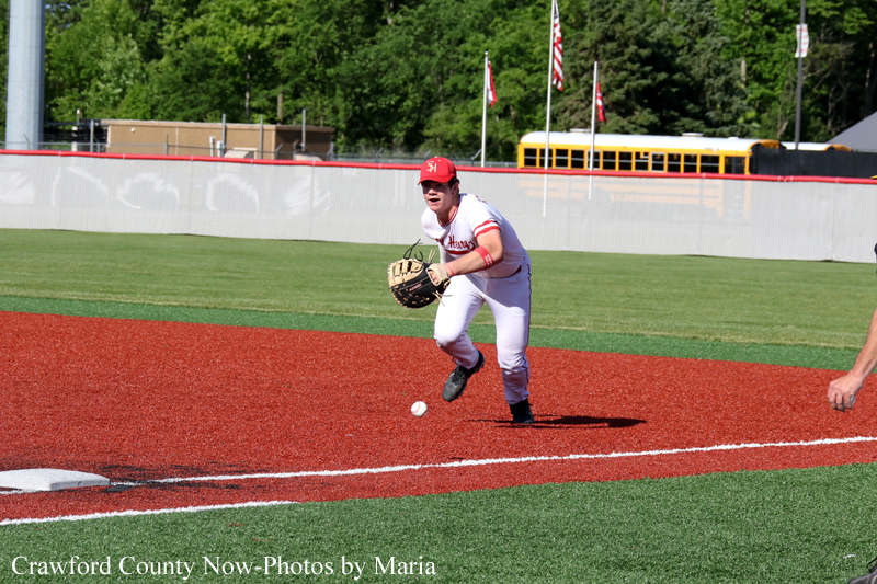 Baseball infielder in white with a red cap fields a ground ball, glove ready, on a red dirt infield under clear skies.