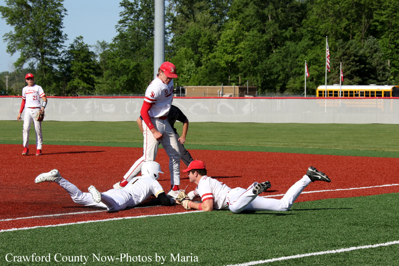 Baseball players in white uniforms slide into home plate as a tag occurs; a teammate stands nearby on a sunny field with a road behind and a bus in the distance.