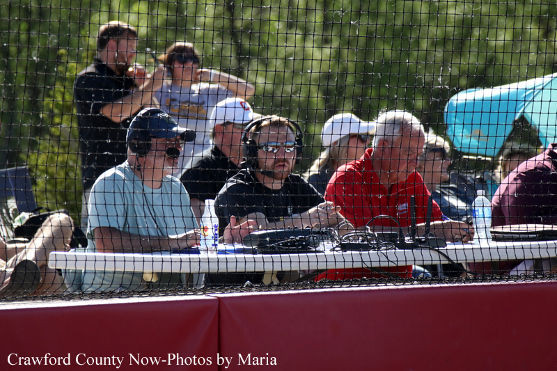 Sports commentators and crew seated at a long table behind protective netting during a game, with headsets and microphones visible.