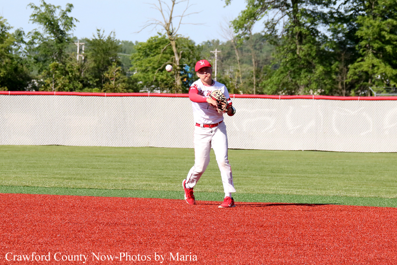 Baseball player in white and red uniform catches a ball on the red dirt infield with the green outfield behind.